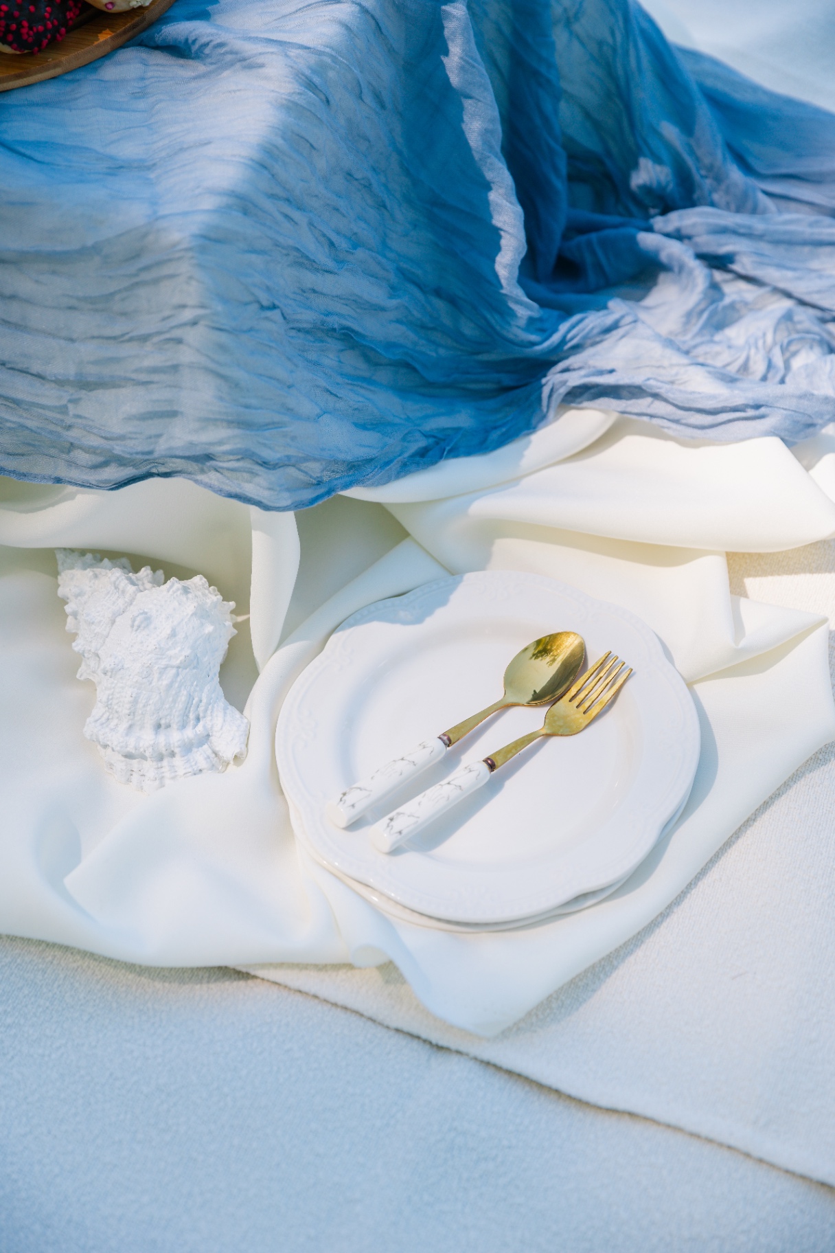 Coastal styling detail with blue gauze runner, white linens, gold cutlery and a seashell