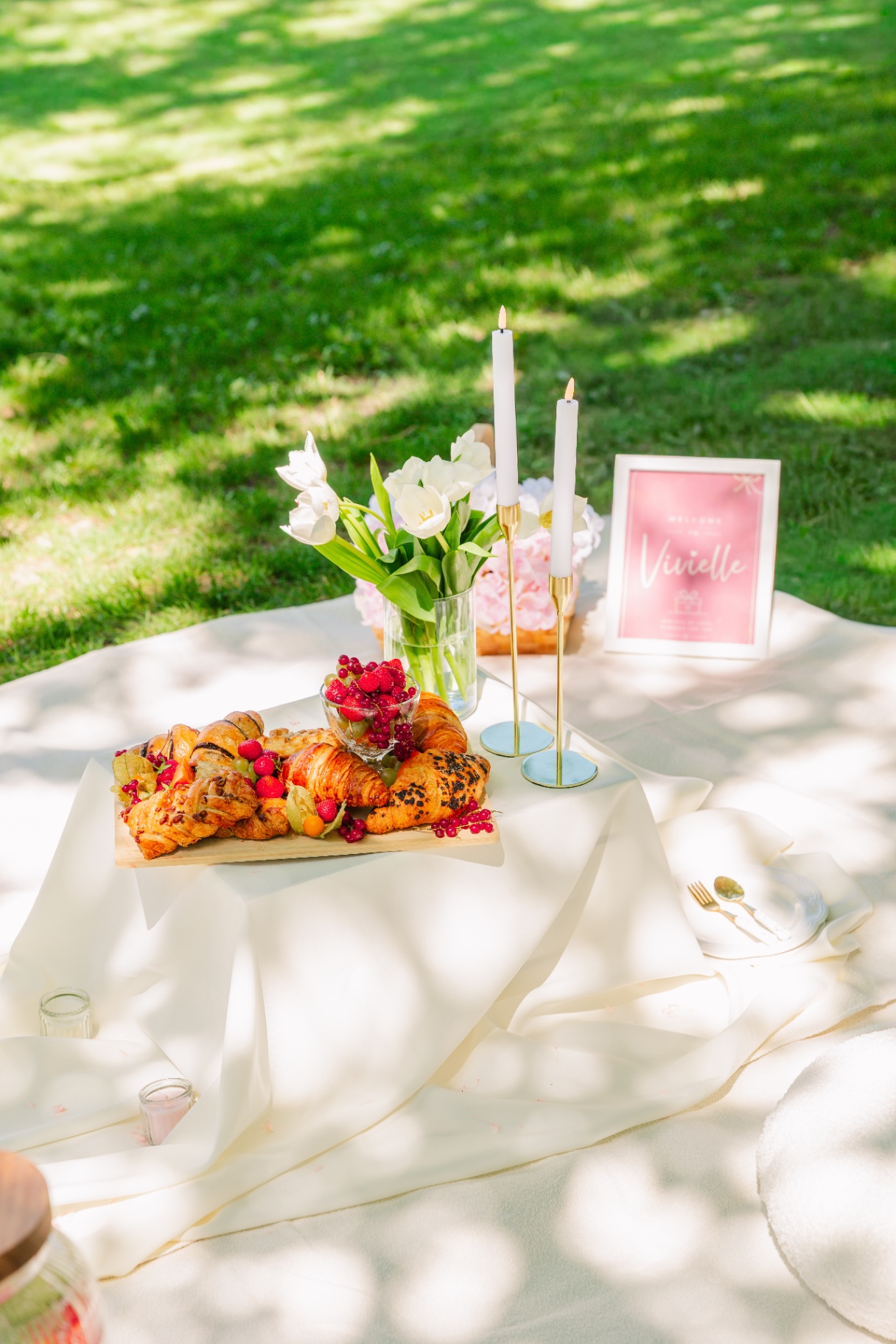 Sunlit picnic with croissant board, white tulips and gold candlesticks on grass