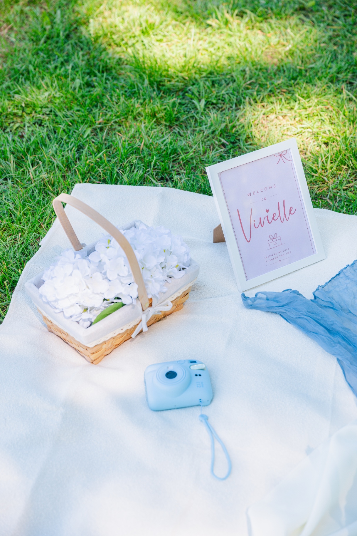 Pastel blue polaroid camera beside a white floral basket on a picnic blanket